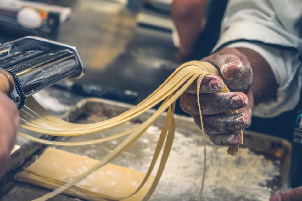 Hands feeding fresh pasta through a roller, dusted with flour